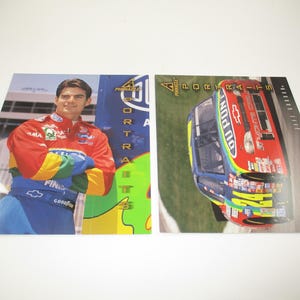 May include: Two trading cards featuring a NASCAR driver and a race car. The driver is wearing a red, blue, and green racing suit and is holding his hands in front of him. The race car is green, blue, and red with the words "Dupont" and "24" on the side.