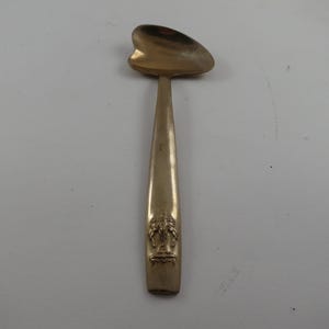 May include: A gold-colored spoon with a heart-shaped bowl and a long, tapered handle. The handle features an embossed design near the base. The spoon is likely made of metal and is set against a plain background.