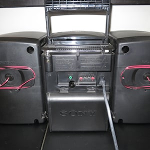 May include: A black Sony boombox with a silver battery compartment cover. The speakers have red wiring visible. The radio antenna is extended. The boombox is on a black surface.