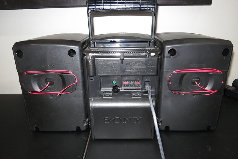 May include: A black Sony boombox with a silver battery compartment cover. The speakers have red wiring visible. The radio antenna is extended. The boombox is on a black surface.