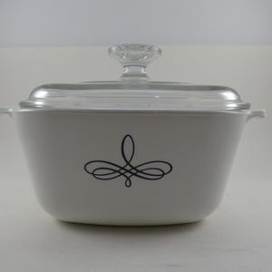 May include: White casserole dish with a clear glass lid and a decorative black swirl design. The dish has two small handles and a clear glass knob on the lid. A classic kitchenware item.