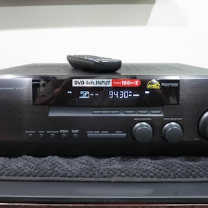 May include: A black Kenwood audio receiver with a remote control on top. The receiver has a digital display showing numbers and text, with various buttons and knobs for volume and input selection. The front panel reads "DVD 6ch.INPUT POWER 100x5".