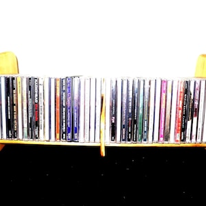 May include: A wooden CD rack with a curved design, holding a collection of CDs. The rack is made of light-colored wood and has a simple, functional design. The CDs are in their jewel cases, with various album covers visible.