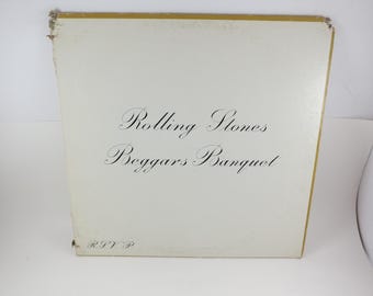 May include: Vintage album cover for The Rolling Stones' "Beggars Banquet." The cover is off-white with gold trim and features the album title and band name in elegant black script. The cover shows signs of wear.