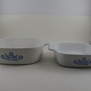May include: Two white ceramic baking dishes with blue floral designs. The dishes have handles and are square-shaped. The larger dish is on the left, and the smaller dish is on the right. The dishes are empty.