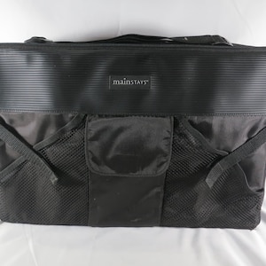 May include: A black rectangular storage organizer bag featuring a horizontal ribbed top panel with a Mainstays logo patch, a central flap pocket, and two side mesh pockets with adjustable straps. The bag includes a top handle for carrying.