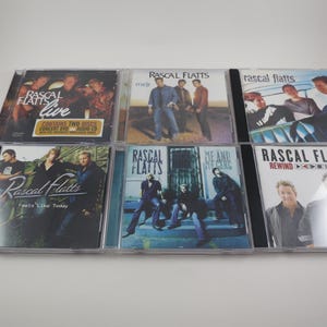 May include: A collection of six Rascal Flatts CDs in clear jewel cases. The album covers feature images of the band members and album titles such as "Live," "Melt," and "Feels Like Today." The CDs are arranged on a white surface.