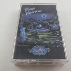 May include: A cassette tape featuring the album art for "Awaken the Guardian" by Fates Warning. The artwork depicts a dark, fantastical landscape with a celestial body and the album title in a stylized font. The cassette is in a clear plastic case.