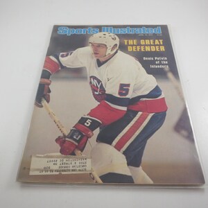 May include: A Sports Illustrated magazine cover from April 18, 1977, featuring hockey player Denis Potvin of the New York Islanders. The athlete wears a white, blue, and orange jersey with the number 5, holding a hockey stick on the ice.