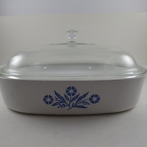 May include: A rectangular, off-white casserole dish with a clear glass lid. The dish features a blue floral design on the front. The lid has a clear glass knob. The dish is approximately 13 inches long.