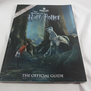 May include: A softcover book titled The Making of Harry Potter, The Official Guide, featuring a New Edition banner. The cover displays a dark forest scene with a person in a red sweater and plaid skirt standing near a large, winged creature.