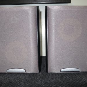 May include: Two gray speakers with a textured surface. Each speaker has a silver accent at the bottom with the brand name. The speakers are rectangular and have a dark border.