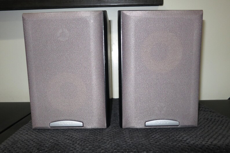 May include: Two gray speakers with a textured surface. Each speaker has a silver accent at the bottom with the brand name. The speakers are rectangular and have a dark border.