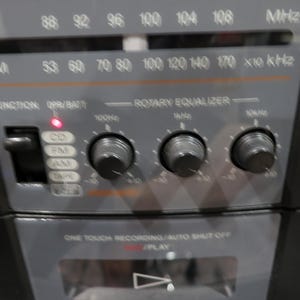 May include: Close-up of a vintage black radio with silver accents. The radio features an FM/AM tuner, a rotary equalizer, and function buttons for CD, FM, AM, and tape. The radio has a red power indicator light and a play button.