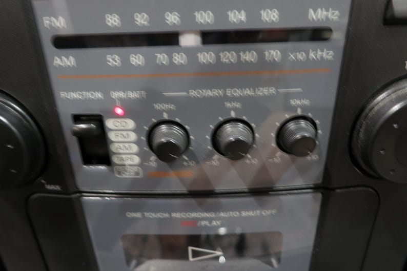 May include: Close-up of a vintage black radio with silver accents. The radio features an FM/AM tuner, a rotary equalizer, and function buttons for CD, FM, AM, and tape. The radio has a red power indicator light and a play button.