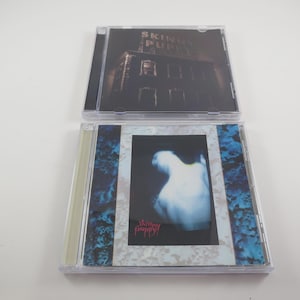 May include: Two CD cases. The top case has a dark cover with the band name "Skinny Puppy" and a building image. The bottom case has a blue and white design with a blurred image and the band name "Skinny Puppy" in pink.