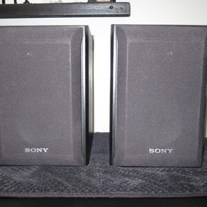 May include: A pair of rectangular black Sony speakers featuring a textured fabric grille covering the front panels. Each speaker has the Sony brand name printed in white lettering at the bottom center of the front face, with a solid black frame.