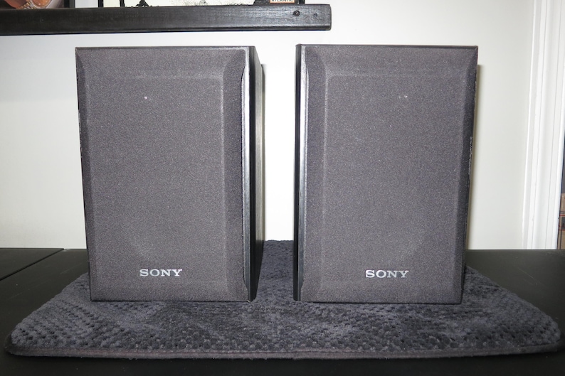 May include: A pair of rectangular black Sony speakers featuring a textured fabric grille covering the front panels. Each speaker has the Sony brand name printed in white lettering at the bottom center of the front face, with a solid black frame.