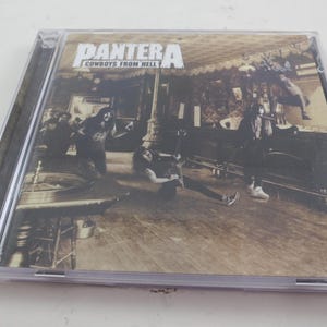May include: A CD case featuring the album "Cowboys from Hell" by Pantera. The cover art depicts a sepia-toned image of a band performing in a saloon. The band's name is in bold white letters at the top.