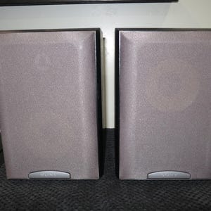 May include: Two gray Sony speakers with a textured surface and black trim. The speakers are rectangular with a slightly angled design. A silver Sony logo is visible at the bottom of each speaker. They are placed on a dark gray surface.