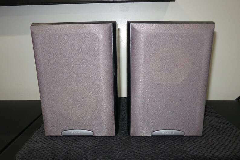 May include: Two gray Sony speakers with a textured surface and black trim. The speakers are rectangular with a slightly angled design. A silver Sony logo is visible at the bottom of each speaker. They are placed on a dark gray surface.