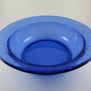 May include: A round, cobalt blue glass bowl with a wide rim. The rim features a delicate, embossed floral pattern. The bowl's interior is a deep, glossy blue, creating a striking contrast with the lighter background.