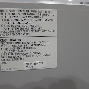 May include: A close-up of a gray device with a white sticker containing text about FCC rules, certification, and manufacturing details. The sticker includes the date "SEPTEMBER 2004".