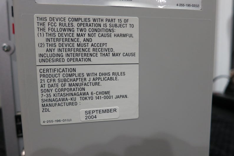 May include: A close-up of a gray device with a white sticker containing text about FCC rules, certification, and manufacturing details. The sticker includes the date "SEPTEMBER 2004".