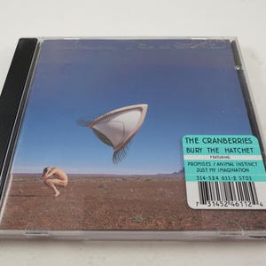Puede incluir: Una caja de CD con una imagen azul y blanca de un objeto volador y una persona de pie en un desierto. El texto en la caja dice "The Cranberries Bury The Hatchet Featuring Promises/Animal Instinct Just My Imagination 314-524 611-2 ST01".