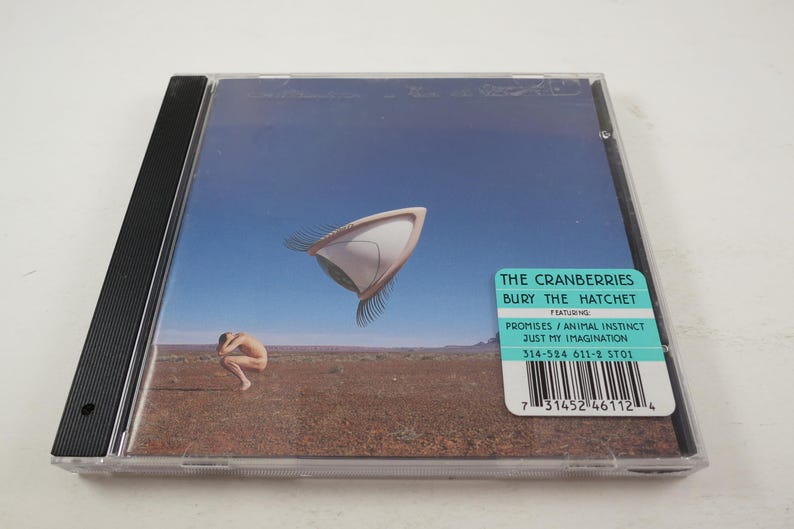 Puede incluir: Una caja de CD con una imagen azul y blanca de un objeto volador y una persona de pie en un desierto. El texto en la caja dice "The Cranberries Bury The Hatchet Featuring Promises/Animal Instinct Just My Imagination 314-524 611-2 ST01".