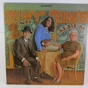 May include: Vintage vinyl record album cover for "Tiny Tim's 2nd Album." The cover features a photograph of three people, with the album title in large, ornate lettering. The background is a mix of green and brown tones.