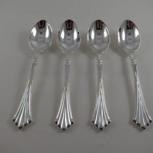 May include: Four silver spoons with a reflective surface are arranged on a light gray background. The spoons have a teardrop-shaped bowl and a decorative handle with a fan-like design.