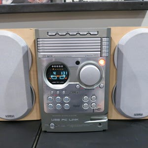 May include: A silver and wood-tone Philips stereo system with two gray speakers. The system has a digital display showing the time and a USB PC Link port. The speakers are mounted on wooden panels.