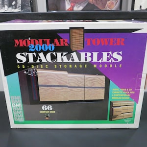 May include: A box for "Modular 2000 Stackables" CD-disc storage module. The box has a black, purple, and teal color scheme. The front of the box features a wooden-look storage unit. The box is designed to hold 66 compact discs.