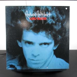 May include: A square album cover featuring Lou Reed with blue and black tones. The album title "Rock and Roll Heart" is in white, and "LOU REED" is in red. The image is mounted on a black base.