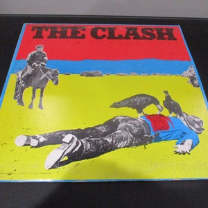 May include: Album cover for "The Clash" featuring a graphic illustration. The top section has red and blue bands, with the band's name in bold letters. The lower section shows a figure lying on a yellow field, with birds and a rider on a horse.
