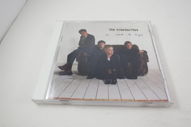 Puede incluir: Una caja de CD con una foto de la banda The Cranberries en la parte delantera. Los miembros de la banda est&aacute;n sentados en un sof&aacute; y el texto "no need to argue" est&aacute; impreso en la parte delantera de la caja.