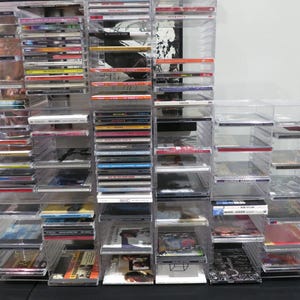May include: Clear plastic CD storage units filled with various CDs. The units are stacked to create a large display. The CDs have colorful covers and spines, creating a visually diverse collection. The storage units are on a black surface.