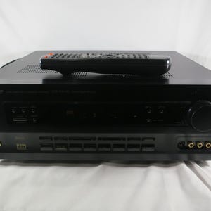 May include: A black Nakamichi AV receiver with a matching black remote control resting on top. The front panel features a large volume dial, multiple input buttons, a headphone jack, and gold-plated audio-video ports with the dts logo.