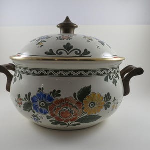 May include: A white ceramic Dutch oven with a lid, adorned with colorful floral patterns in blue, orange, and yellow. The pot has dark brown handles and a gold rim, perfect for cooking and serving.