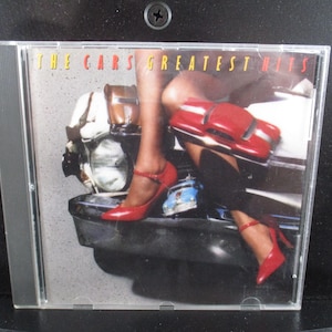 May include: A red and black CD case with the text "The Cars Greatest Hits" on the front. The case features a woman wearing red shoes and a red car in the background.