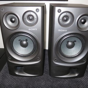 May include: A pair of black Sony bookshelf speakers featuring a two-way design with a large woofer, a smaller tweeter, and a bass reflex port at the bottom. The Sony logo is printed in white on the front of each rectangular speaker cabinet.