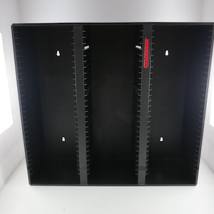 May include: A black storage case with multiple vertical dividers. The case has a rectangular shape and is designed to hold items. The dividers are evenly spaced, and there is a red label on the side. The case is empty.