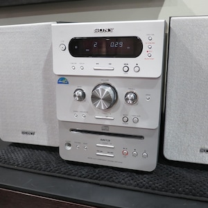 May include: A silver and white Sony stereo system with two speakers. The stereo has a digital display, CD player, and various control buttons. The speakers are rectangular and have the Sony logo.