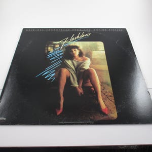 May include: A black vinyl record album cover with the title "Flashdance" in blue and white text. The cover features a woman wearing a white shirt and red shoes, sitting on a bench.