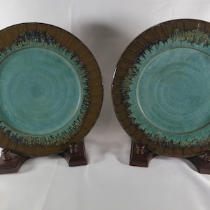 May include: Two round ceramic plates featuring a smooth, light blue center and a wide, dark brown rim. The transition between the blue and brown sections is marked by a textured, mottled band of dark green and black glaze with a radial pattern.