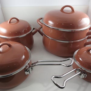 May include: A set of brown enamel cookware with lids and silver-colored handles. The set includes various sized pots and pans, arranged in a white box. The pots have a rounded shape and a shiny finish, suitable for cooking.