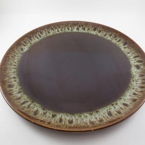 May include: A round, ceramic plate with a dark brown center and a textured, light green and brown rim. The plate has a glossy finish and is set against a white background. This plate is suitable for serving food.