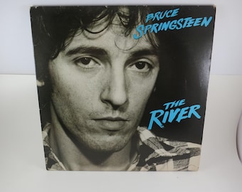 May include: Album cover for Bruce Springsteen's "The River." The black and white image features a close-up of Springsteen's face. The album title and artist's name are in blue text. The cover has a vintage aesthetic.