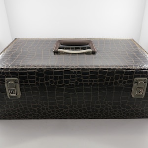 May include: A dark brown, rectangular storage box with a faux crocodile skin texture. The box has a brown handle and two silver latches. The box is sitting on a white surface.